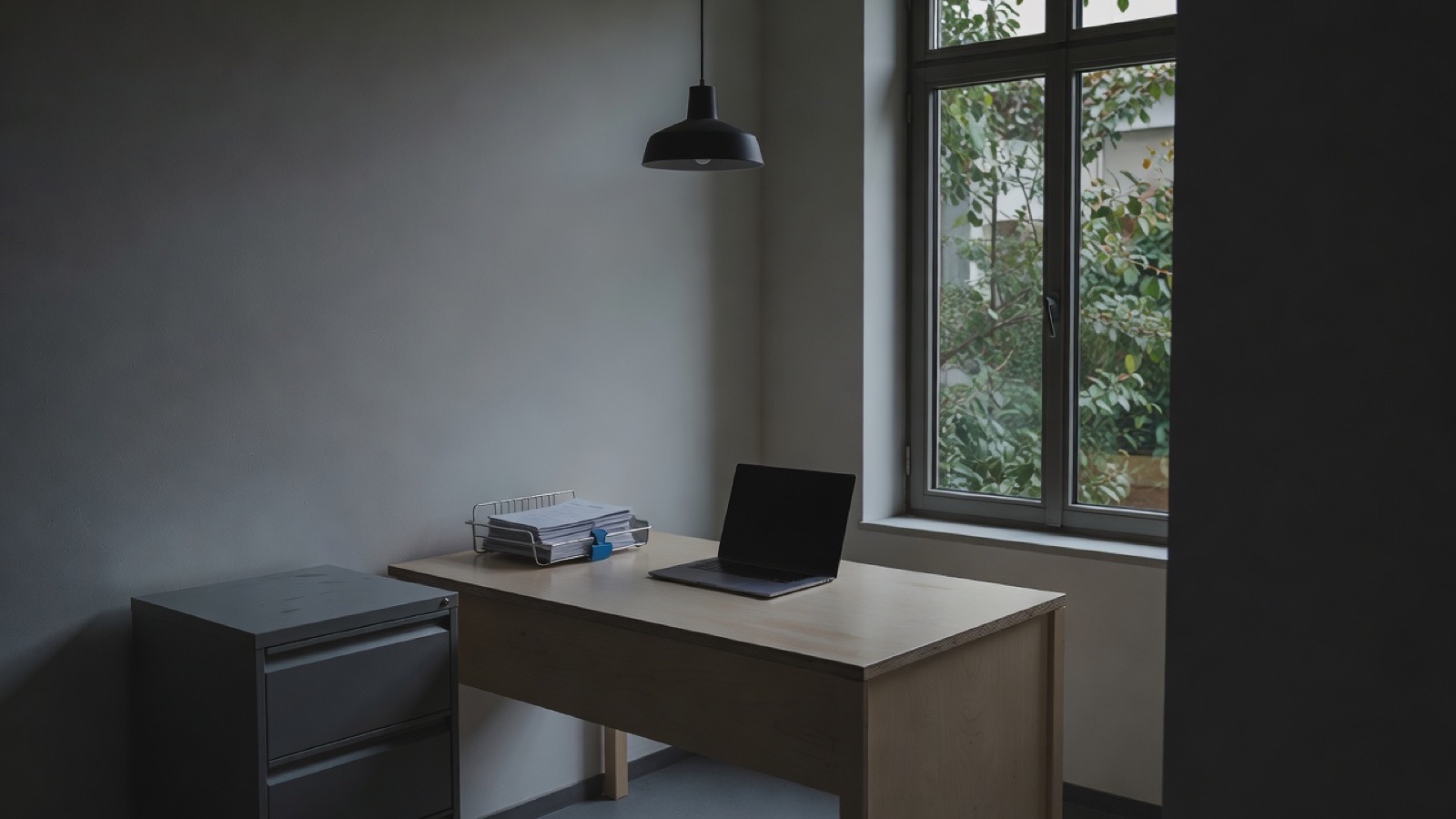 A quiet back office in the early evening: a closed laptop on a pale desk, a paper invoice tray, a filing cabinet and a window onto a courtyard