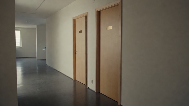 Two identical office doors side by side in a quiet corridor, one ajar, with a small brass plate on each and light from a window at the end